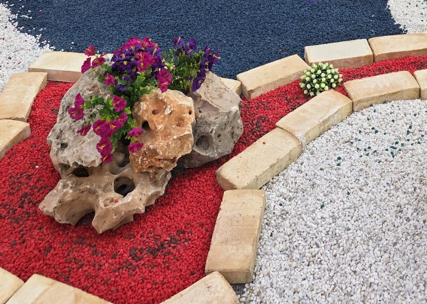 Jardín decorativo con piedra blanca, grava coloreada roja y negra, borduras de piedra y plantas ornamentales, creando contrastes de color en paisajismo exterior.